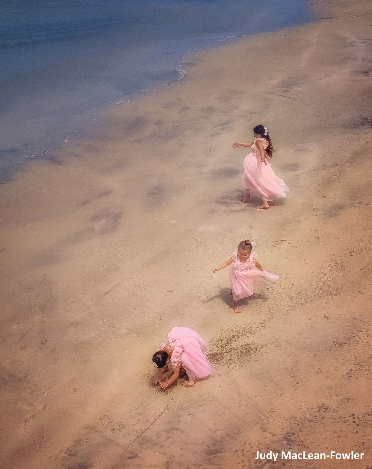 Three young childen in pink dresses dance around on the beach.